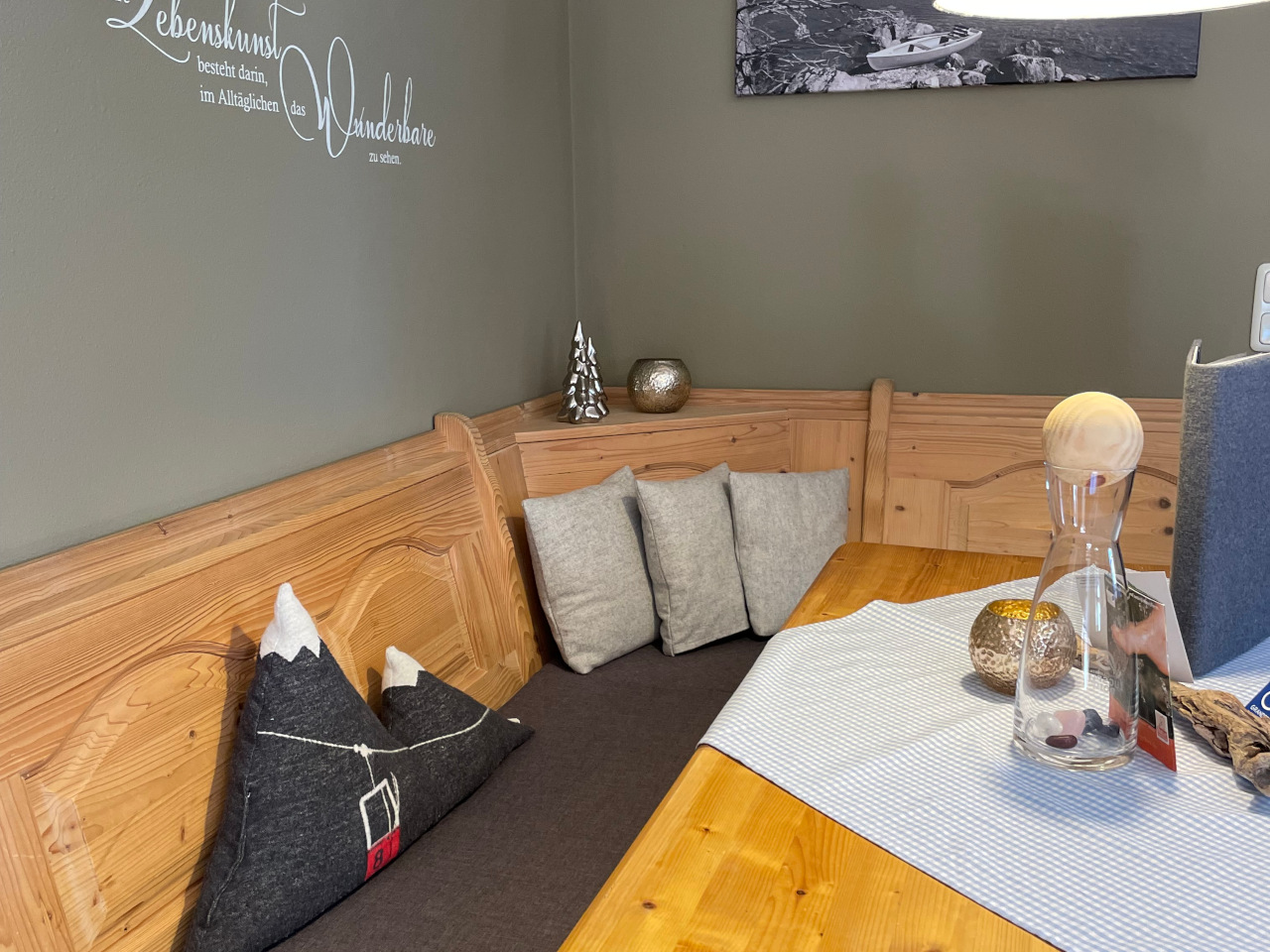 gemütliche Eckbank aus Holz mit einem Kissen mit Gondel, braun-grauer Wand mit einem Spruch und einem Bild von einem See, auf dem Tisch mit einer karierten Decke steht ein Wasserkrug