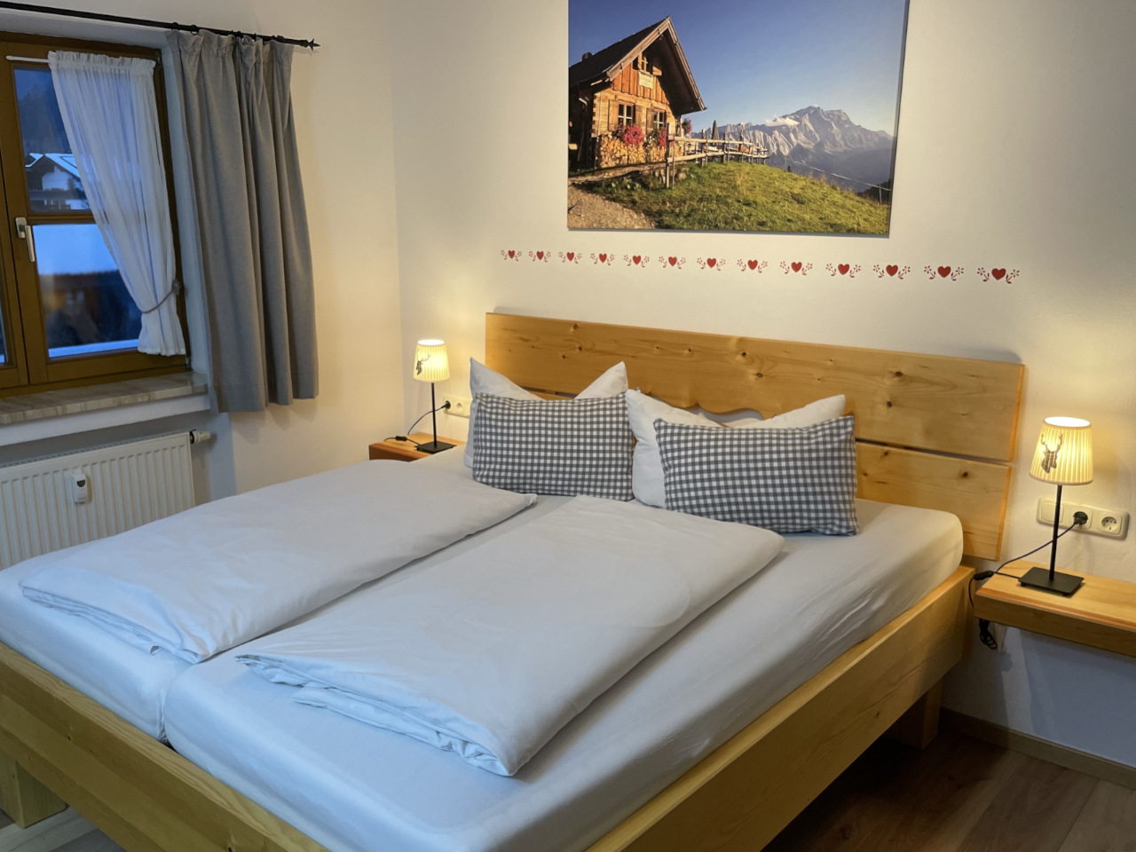 Gemütliches Schlafzimmer mit Doppelbett aus hellem Holz, weißer Bettwäsche und karierten Kissen; links ein Fenster mit Vorhängen, rechts und links Nachttische mit kleinen Lampen; über dem Bett hängt ein Bild einer Berghütte in alpiner Landschaft.